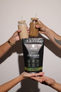 Hands holding a front-facing Origin Collagen 250g collagen pouch with two iced coffee glasses clinking above, showcasing a lifestyle moment with the product